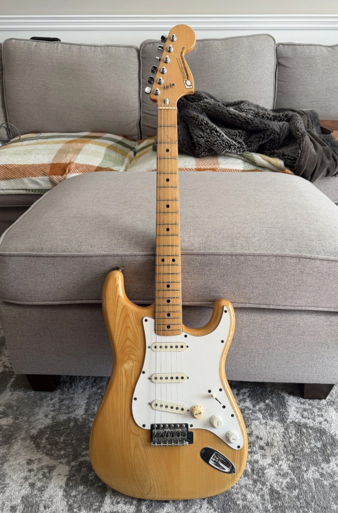 A close-up view of an electric guitar, featuring a natural wood finish, white pickguard, and three pickups, placed on a light gray sofa.