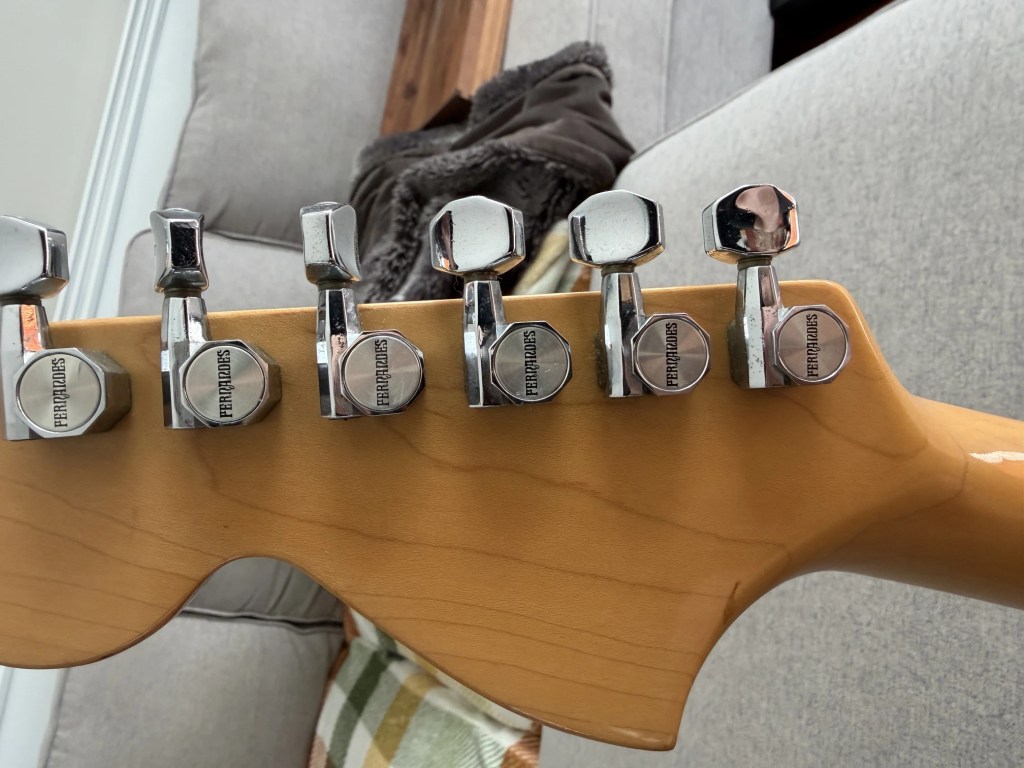 Close-up view of the headstock of a guitar, showcasing the tuning pegs and part of the neck, with a soft background featuring a couch.