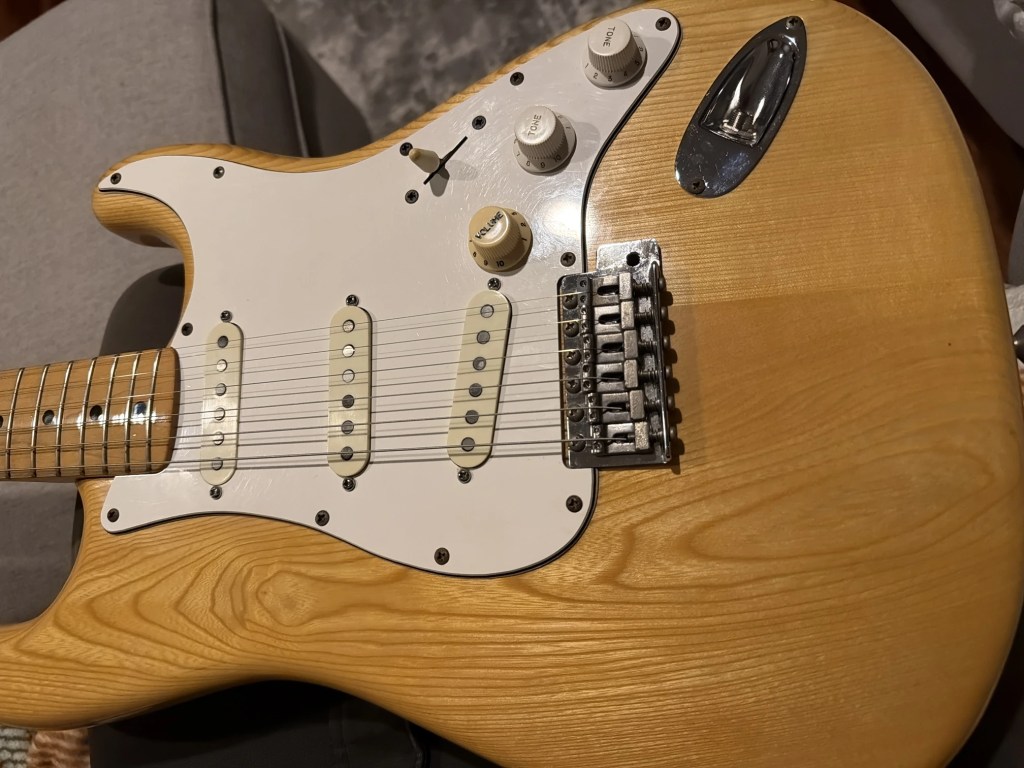 Close-up of an electric guitar featuring a natural wood finish, white pickguard, three single-coil pickups, and control knobs for volume and tone.