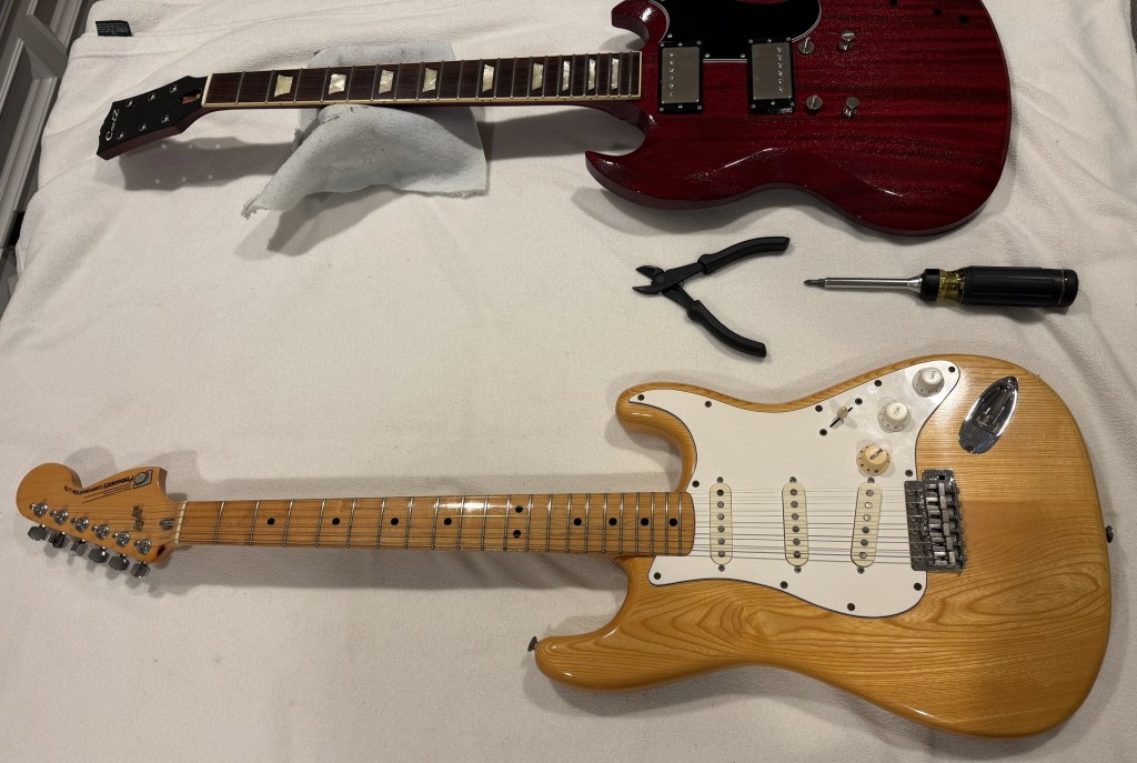 A top view of two electric guitars on a fabric surface, one is a red SG model with a solid body, and the other is a natural wood Stratocaster. Nearby tools, including pliers and a screwdriver, are visible.