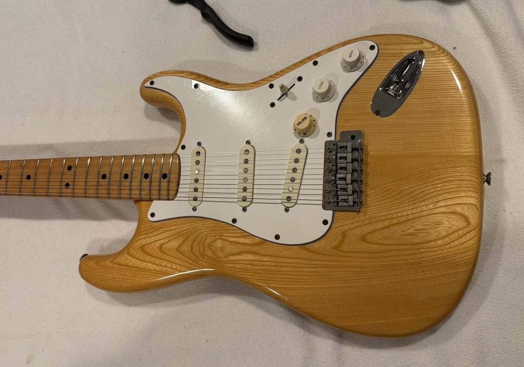 An electric guitar with a natural wood finish, white pickguard, and multiple control knobs, lying on a light surface.