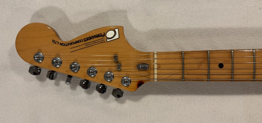 Close-up view of a Fender guitar neck with tuning pegs and frets, displayed on a neutral background.