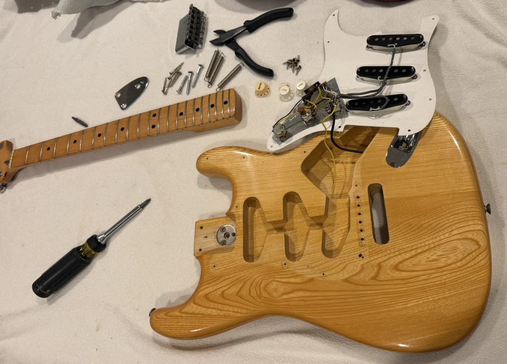 Disassembled electric guitar body with a neck, pickups, and various hardware components laid out on a light-colored surface.
